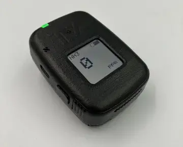 TDA Research’s wearable chemical sensor badge lays on a white, plain surface. The badge’s features include a button on the bottom left-hand side; a digital readout in the front-center that displays information such as battery life, type of toxic industrial chemical present and the amount in parts-per-million; and a green indicator light on its top left-hand side. 