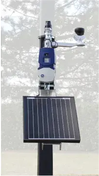 N5 Beta Wildfire Sensor mounted on post. Solar panel at bottom, sensor box in middle, wind sensor at top. Located outdoors with trees and grass behind sensor set up.