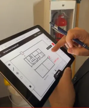 A man’s left hand holds a tablet showing Mappedin floor plan of a building. While his right hand is holding a pen, his finger is touching the tablet’s screen. 