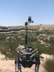 A black 4-legged metallic robot dog is standing in a sandy desert environment. There is dry scrub brush everywhere. A black fence runs all the way to a sandy earthen hilly embankment in the far distance. A metal rod with a camera/sensor package has been deployed from the back of the robot dog to monitor the area.