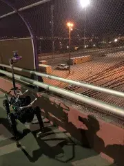 At night, a robot dog is walking across a bridge overpass that is above a railyard. Through the chain link fencing on the bridge, train tracks, a pick-up truck and shipping crates are visible. The robot dog has its camera/sensor package extended up and is surveying the railyard.