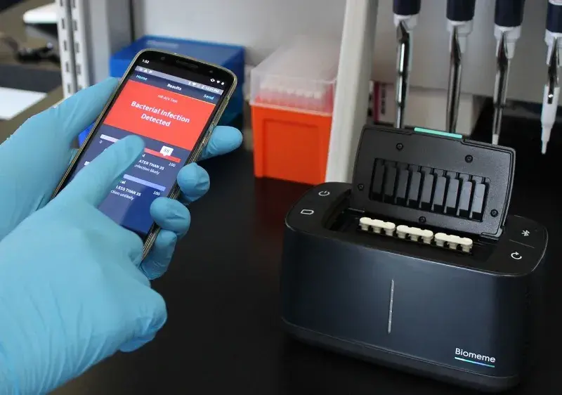 A researcher points to a result reading “Bacterial Infection Detected.” The black Franklin™ thermocycler by Biomeme, Inc. (right) can measure any DNA or RNA that Predigen, Inc. designs a test for. Inside the thermocycler are white test tubes with reagents to detect host response signature in samples. It can be operated by a smartphone app and can export data to the cloud.  Photo by Biomeme/Predigen.